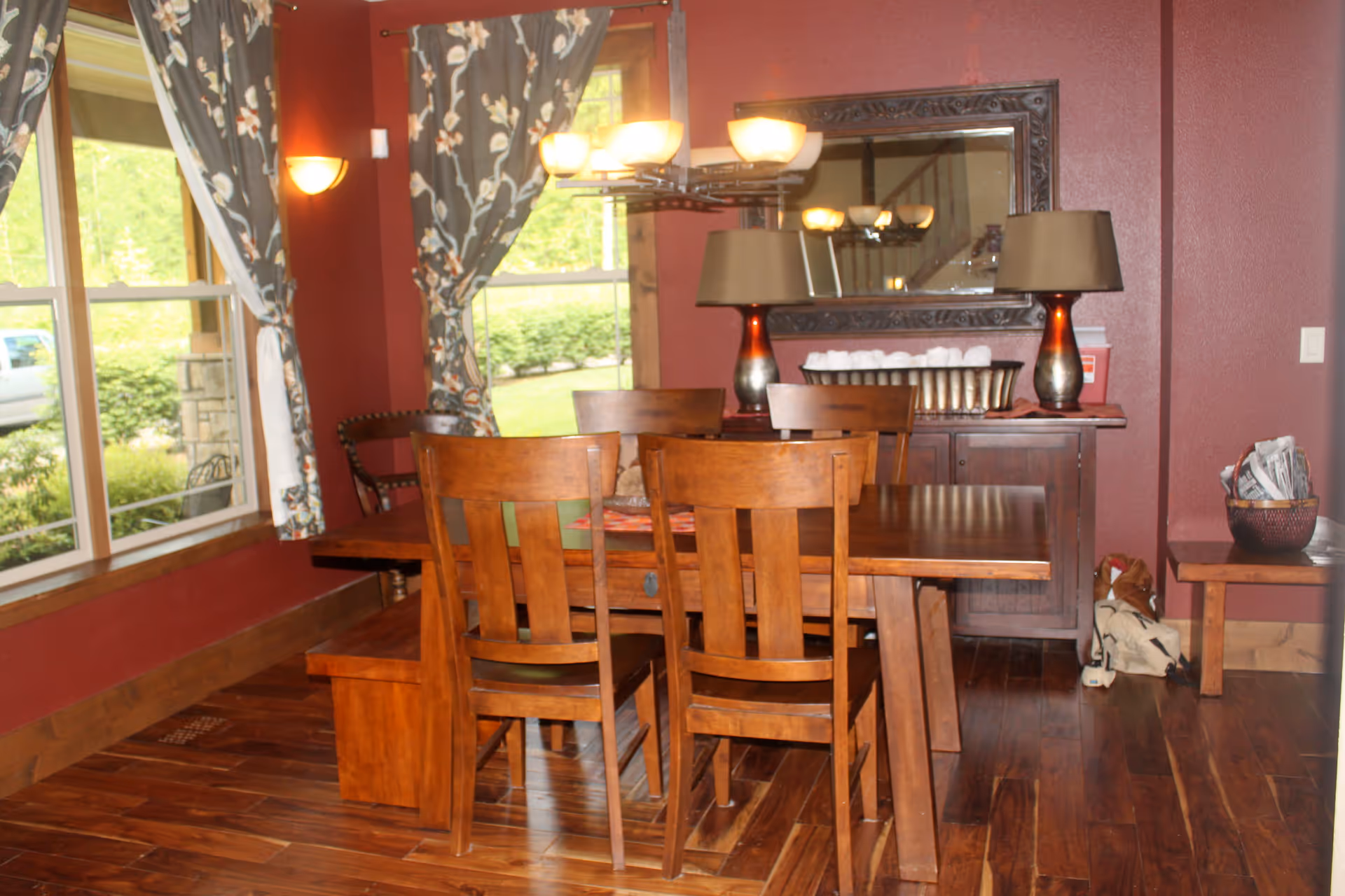 A dining room with a wooden dining table and four wooden chairs. The room has dark red walls, a large window with floral curtains, and a wooden sideboard with two lamps and a decorative mirror above it. There is a basket with newspapers on a small bench to the right and a view of greenery outside the window.