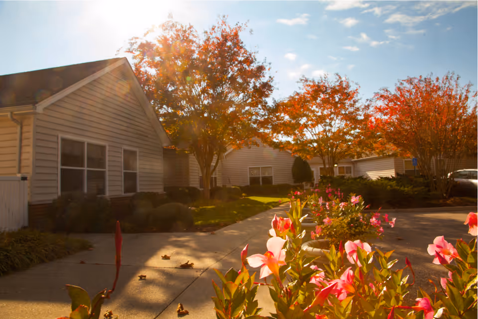 Sunlit courtyard with flowering shrubs and single-story senior care buildings framed by autumn trees.