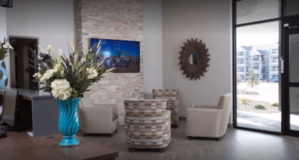 A cozy seating area in a senior living facility with four armchairs arranged around a small table. The room features a stone accent wall with a mounted flat-screen TV, a decorative round mirror on the adjacent wall, and a large window and glass door letting in natural light. A turquoise vase with white and purple flowers is placed on a wooden table in the foreground.