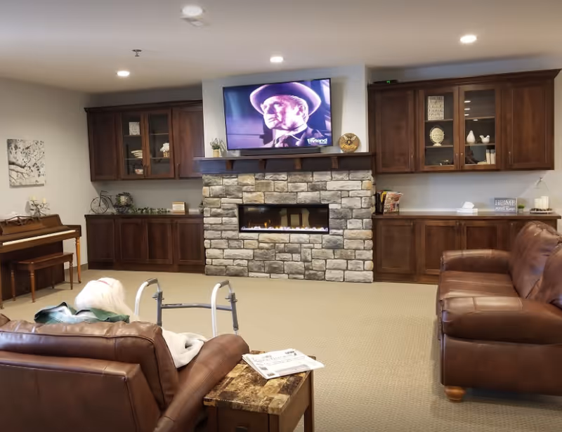 Cozy senior living lounge with stone fireplace, mounted TV, piano, and leather seating.
