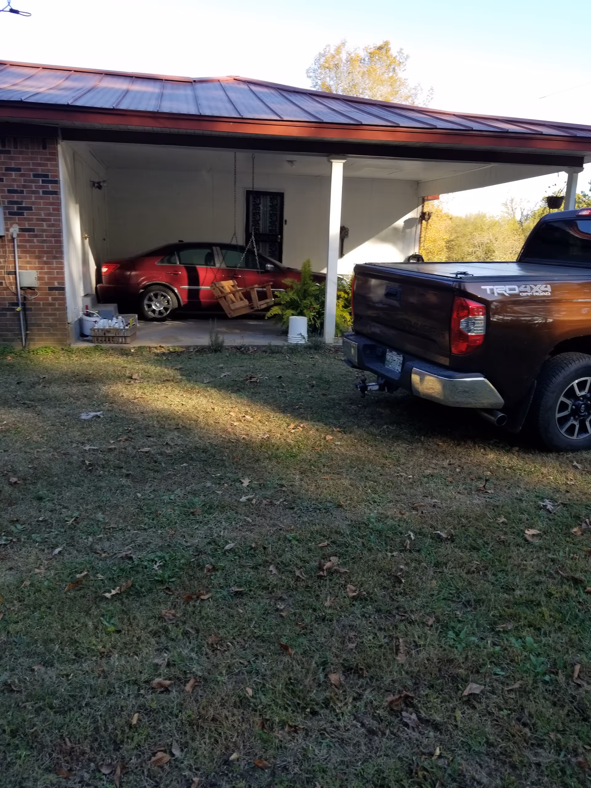 Red car and brown pickup under a covered carport at a residential home.