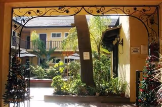 View through an archway into a courtyard garden area with plants, trees, and umbrellas. The courtyard is surrounded by a two-story building with balconies and windows. The archway has decorative wrought iron details.