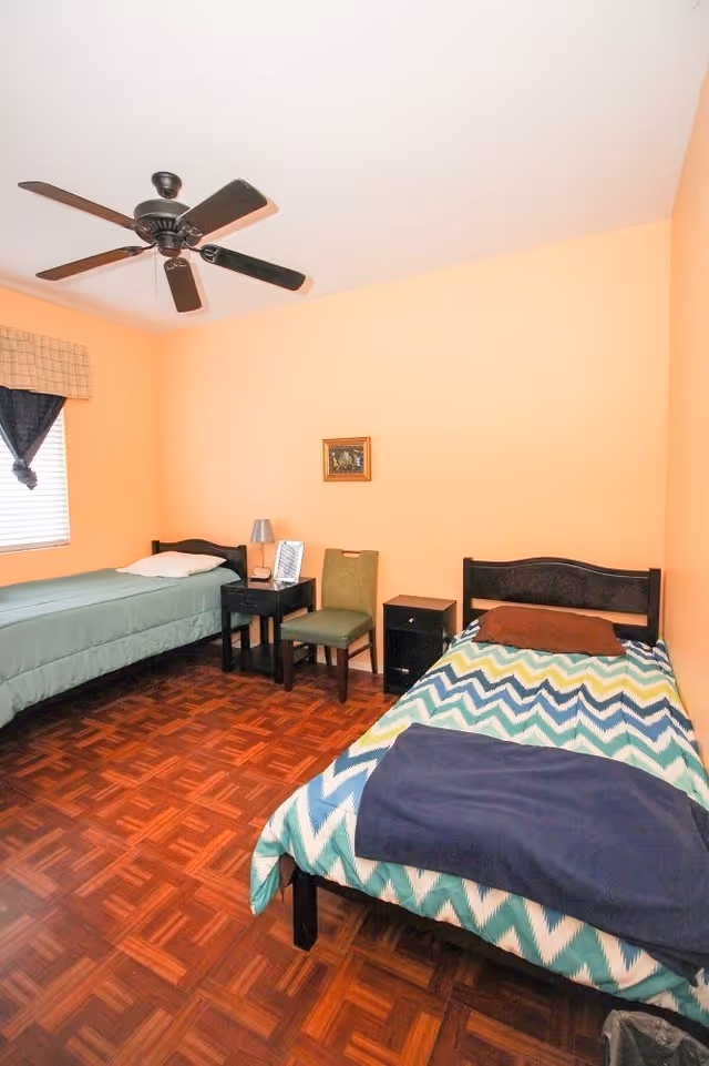 A small bedroom with two single beds, one with a green bedspread and the other with a blue and white zigzag patterned bedspread. There is a green chair and two small black nightstands between the beds, one with a lamp and a framed photo. The walls are painted peach, and there is a ceiling fan with dark blades. A window with a plaid valance and dark curtains is on the left wall.