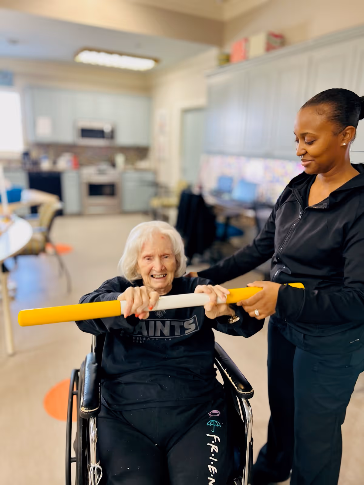 An elderly woman in a wheelchair holding a yellow and white exercise bar with the assistance of a caregiver in a black outfit. They are in a room with a kitchen area in the background, including cabinets, a stove, and a microwave.