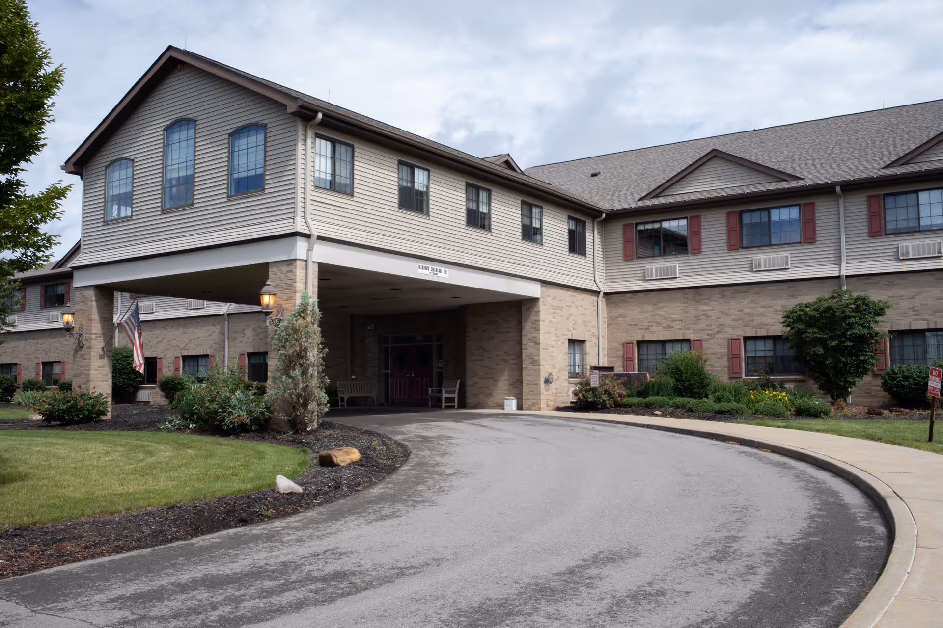 Covered entrance and circular driveway of a two-story brick and siding senior living building with landscaping and an American flag.