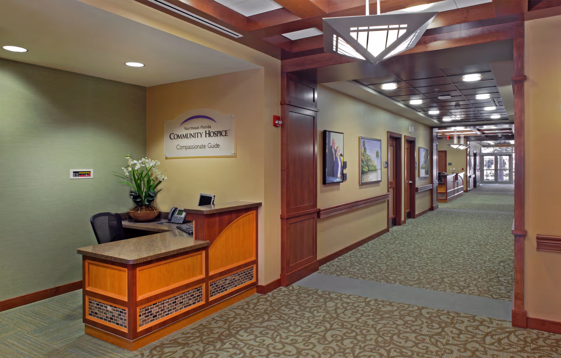Reception desk in a warm hospice corridor with framed art and overhead lighting.