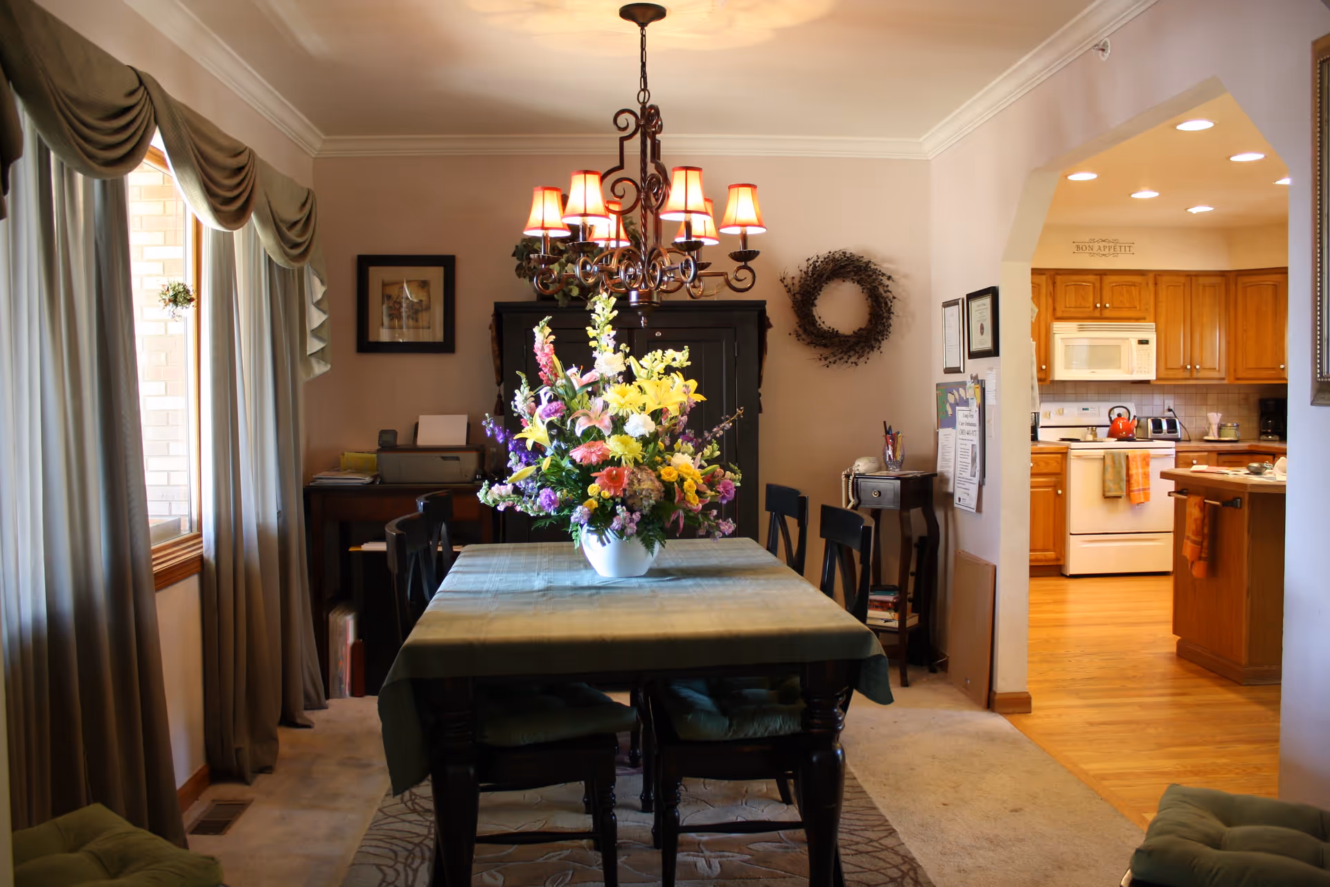 Dining room with a table topped by a large floral centerpiece under a chandelier and a view into a kitchen.