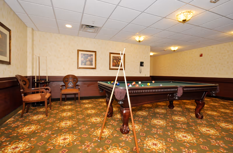 A billiards room with a pool table, cue sticks propped up, chairs, framed artwork, and patterned carpet.