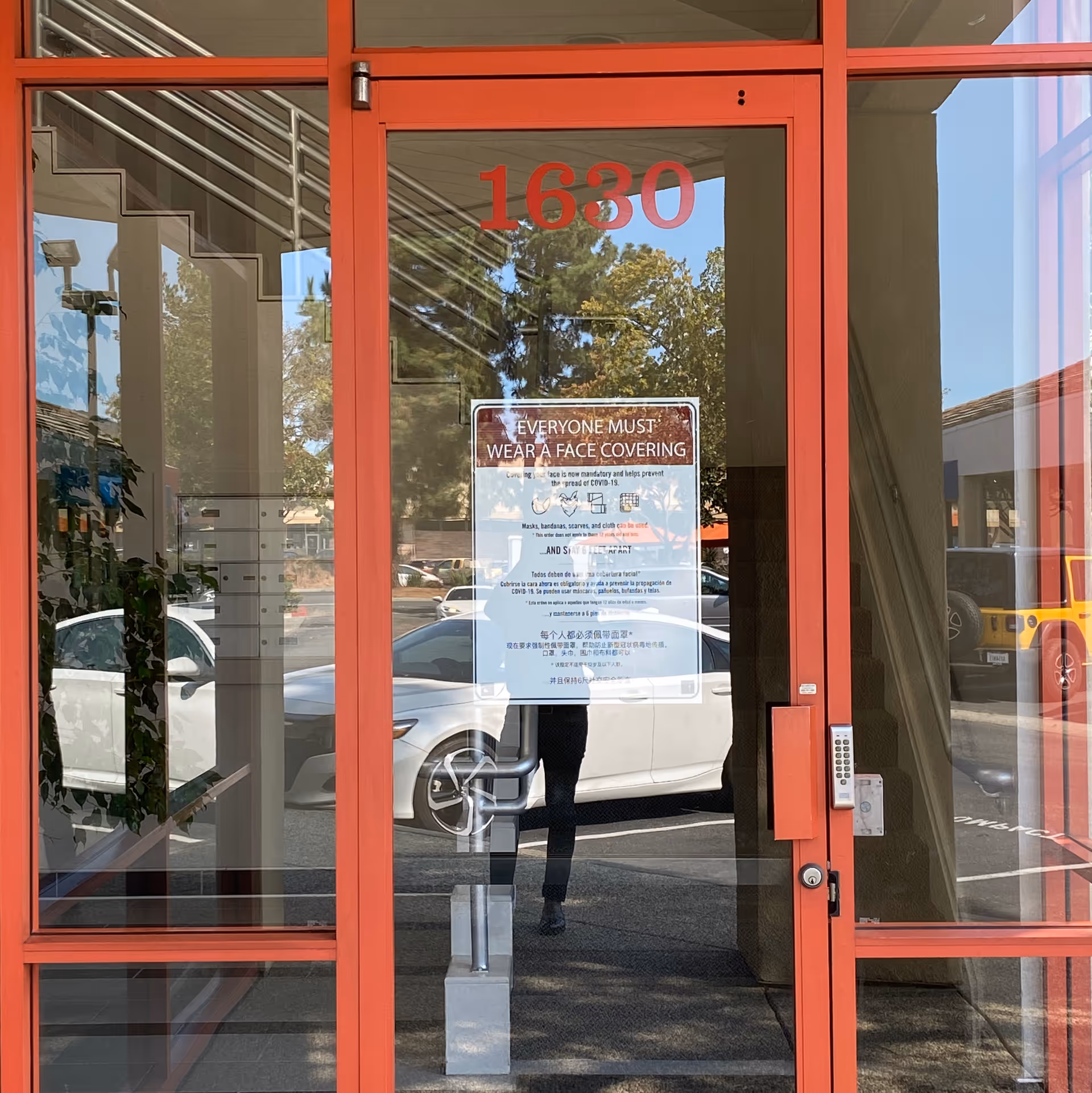 Red-framed glass entrance with a posted face-covering sign and reflections outside.