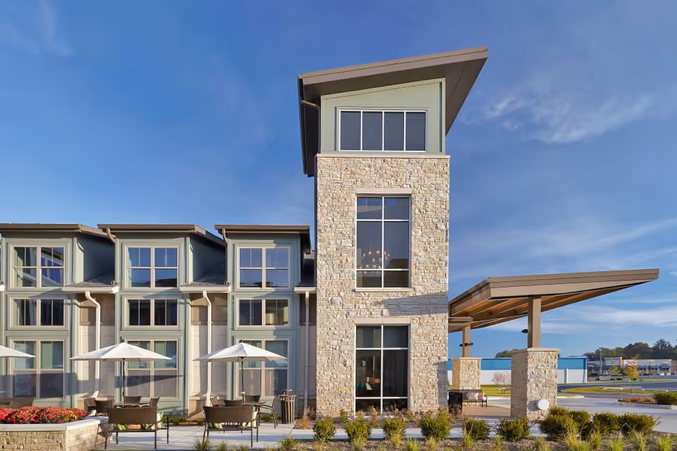 Exterior view of a modern senior living facility building with large windows, stone and light green siding, outdoor seating with umbrellas, and a clear blue sky.