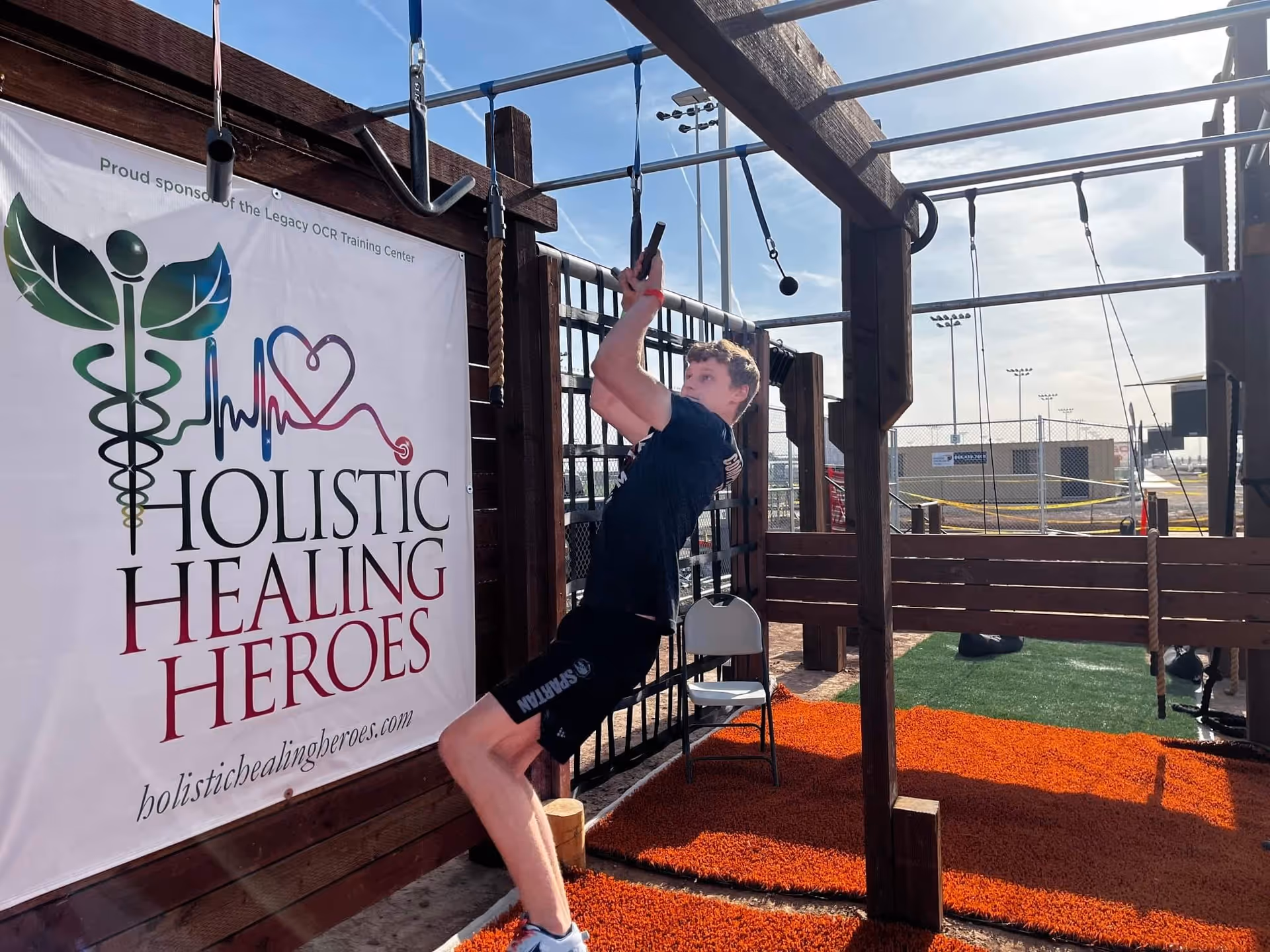 A man climbs an outdoor wooden obstacle course beside a Holistic Healing Heroes banner.