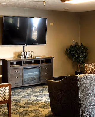 A cozy living room area with a mounted flat-screen TV above a wooden cabinet that has decorative letters 'SVM' on top. There is a patterned carpet on the floor, a potted plant in the corner, and upholstered chairs with patterned fabric.