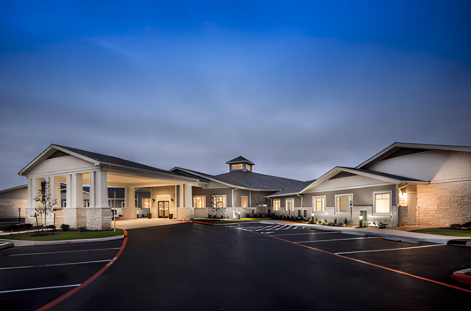 Exterior front entrance of a single-story senior living facility at dusk with a covered porte-cochere and empty parking lot.