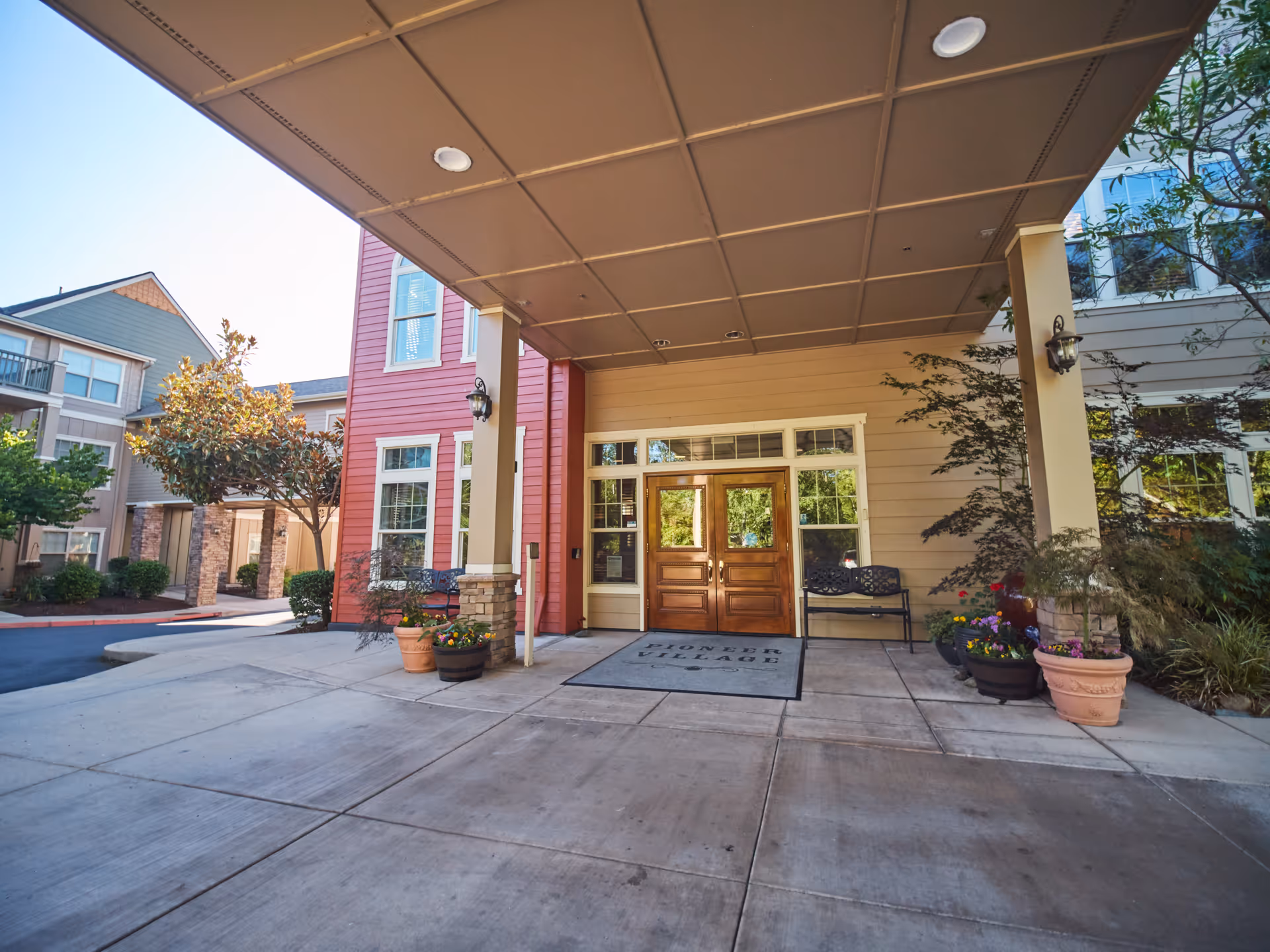 Covered entrance of the Pioneer Village senior living facility with double wooden doors, potted plants, and a bench under a porte-cochere.
