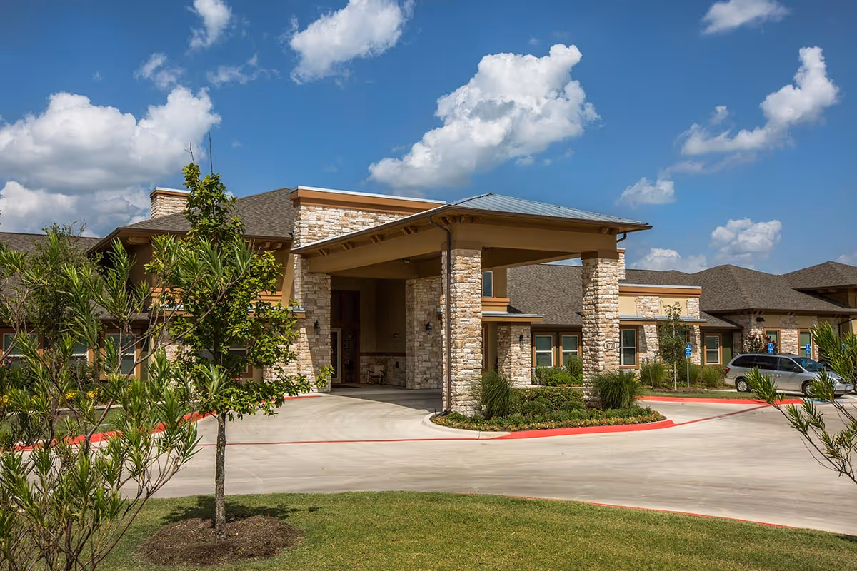 Front entrance of a stone-clad senior living facility with a covered porte-cochere, driveway, and landscaped grounds under a blue sky.