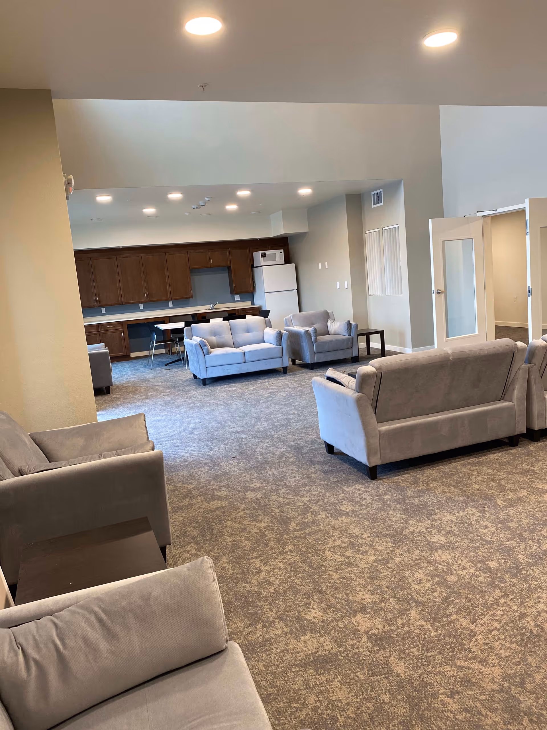Spacious communal lounge with multiple gray sofas, carpeted floor, and a kitchenette with cabinets and a refrigerator in the background.
