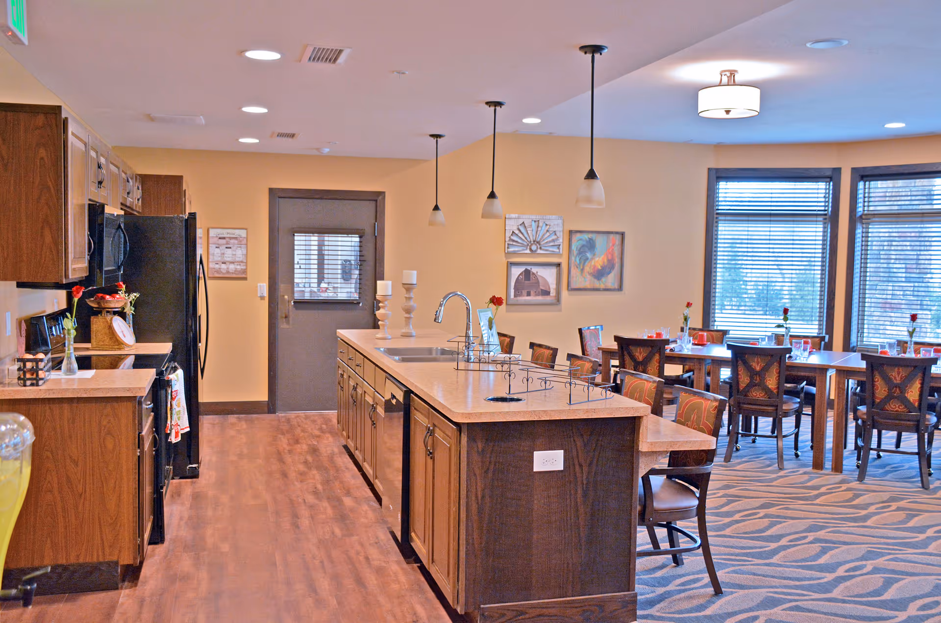 A spacious kitchen and dining area in a senior living facility. The kitchen features wooden cabinets, a black refrigerator, a stove, and a large island with a sink and pendant lights above. The dining area has several tables and chairs, each table decorated with a single red rose in a vase. Large windows with blinds allow natural light to fill the room. The walls are painted a warm beige color and decorated with framed artwork.