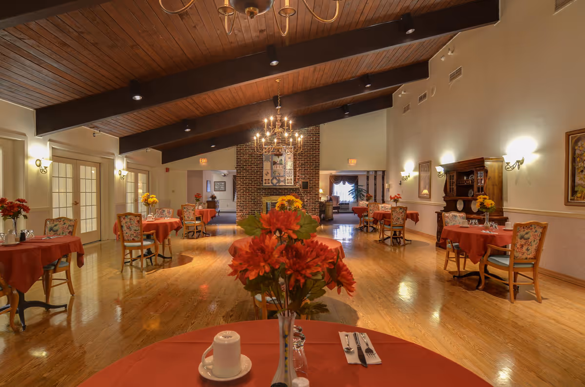Spacious dining room with red-clothed tables, floral centerpieces, wooden chairs, and a brick fireplace.