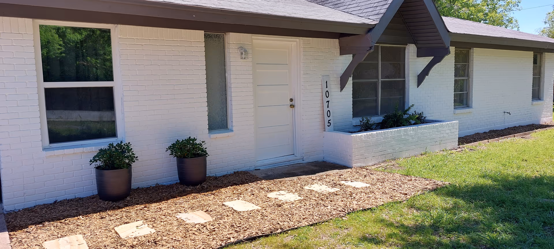 White brick senior living facility exterior with front entrance, address plaque, planters, and stepping stones.