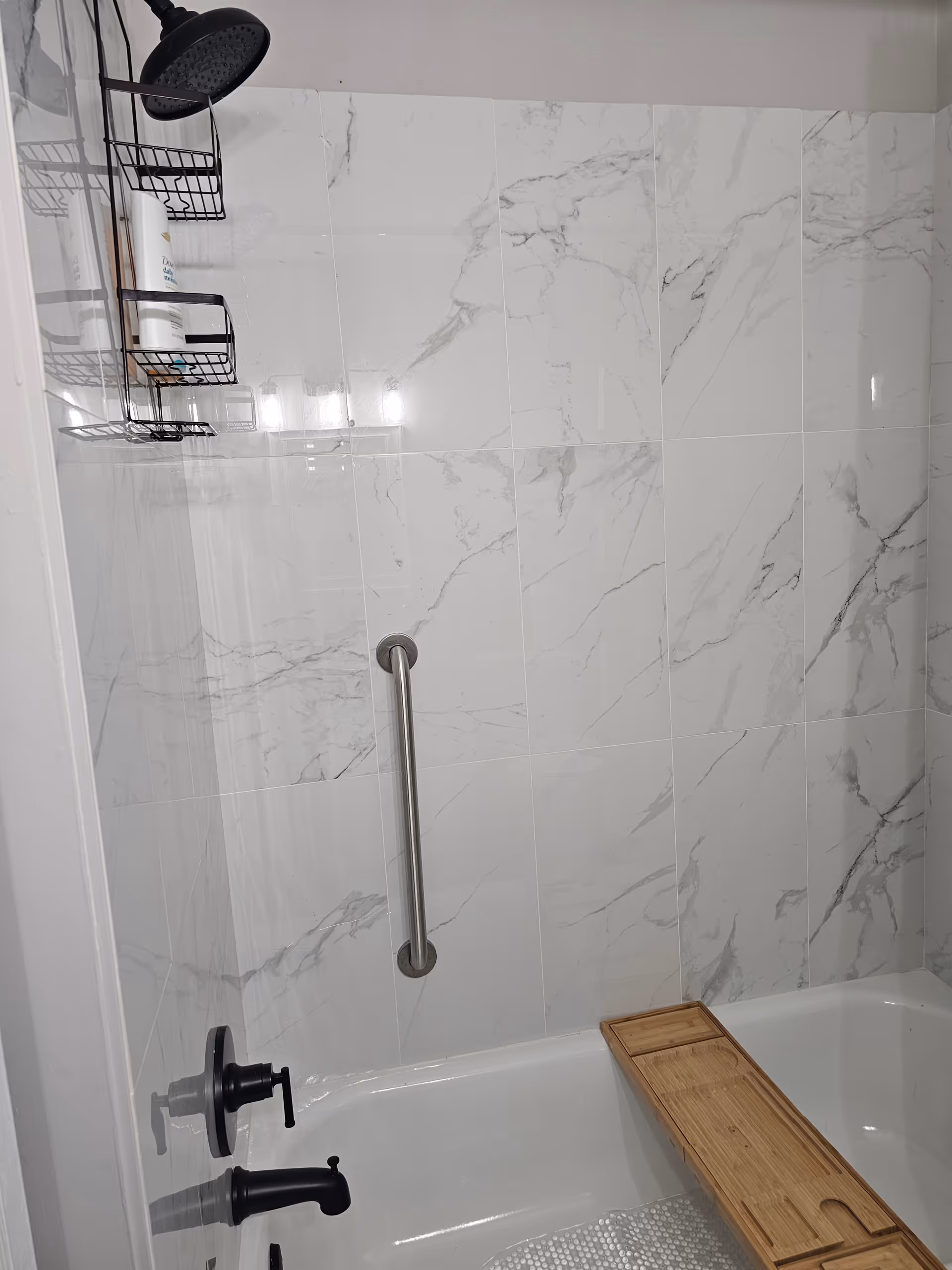 Shower with marble tile walls, a grab bar, and wooden bath board in bathroom.