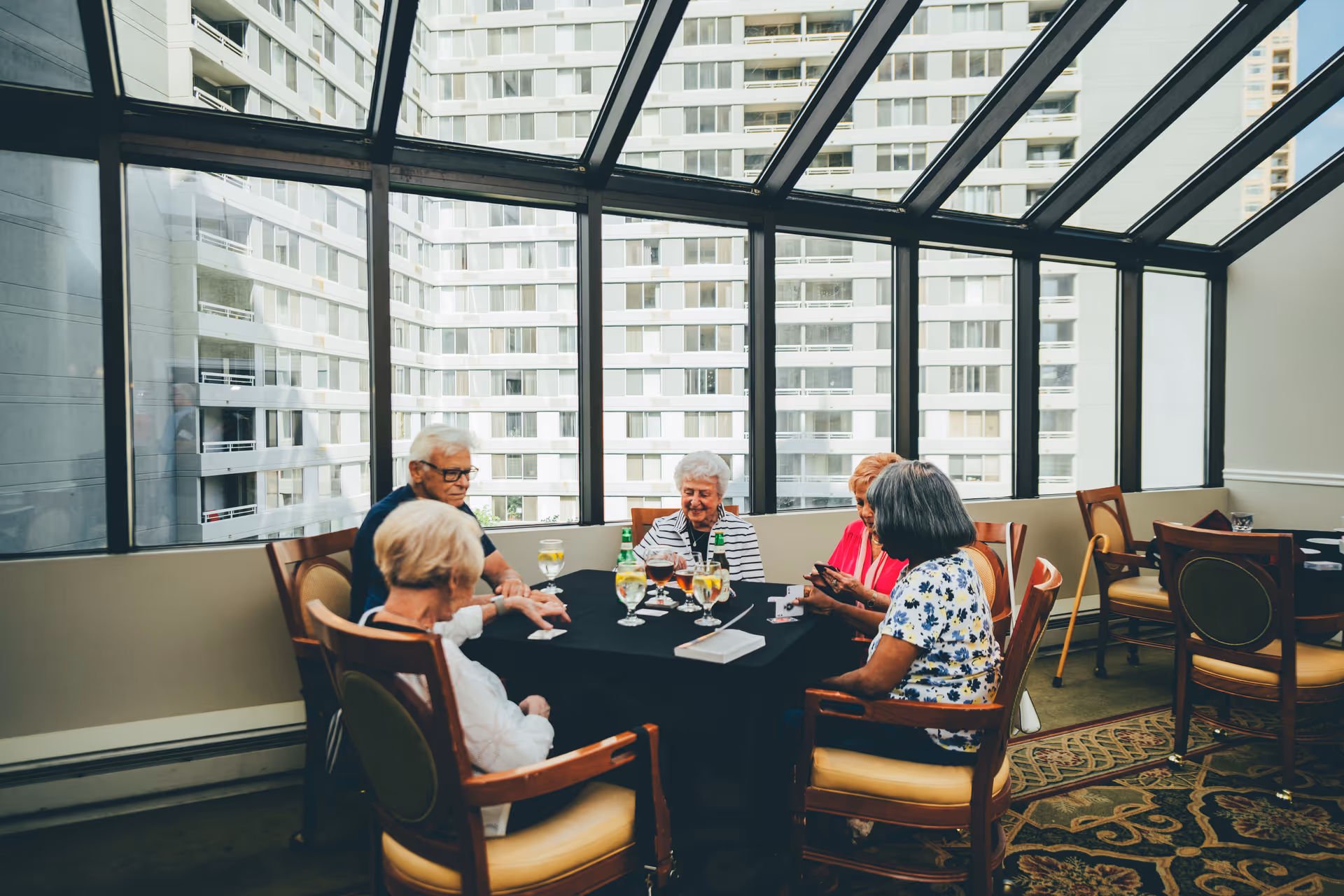 A group of elderly people sitting around a table playing cards in a sunlit room with large windows showing a cityscape outside.