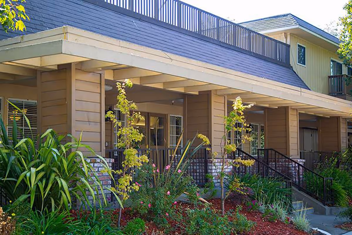 Exterior view of Silverado Berkeley Memory Care Community building entrance with a covered porch, surrounded by landscaped greenery including small trees, shrubs, and flowers.