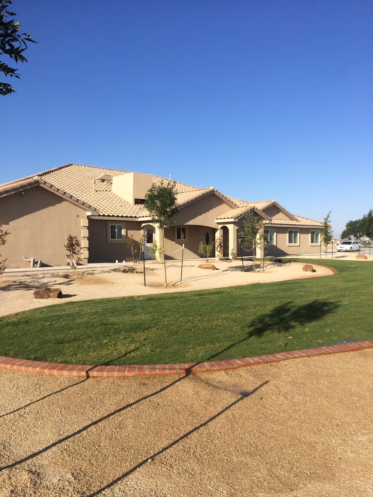 Exterior view of a single-story senior living facility building with beige walls and a tiled roof under a clear blue sky. The foreground shows a landscaped area with green grass, small trees, and a gravel pathway bordered by bricks.
