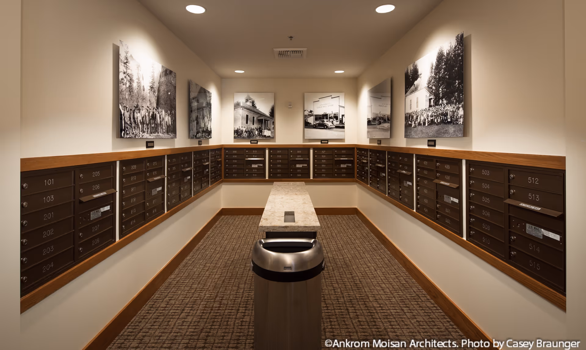 Interior mail room with rows of numbered mailboxes along both walls, a central table and trash can, and framed black-and-white photos above.