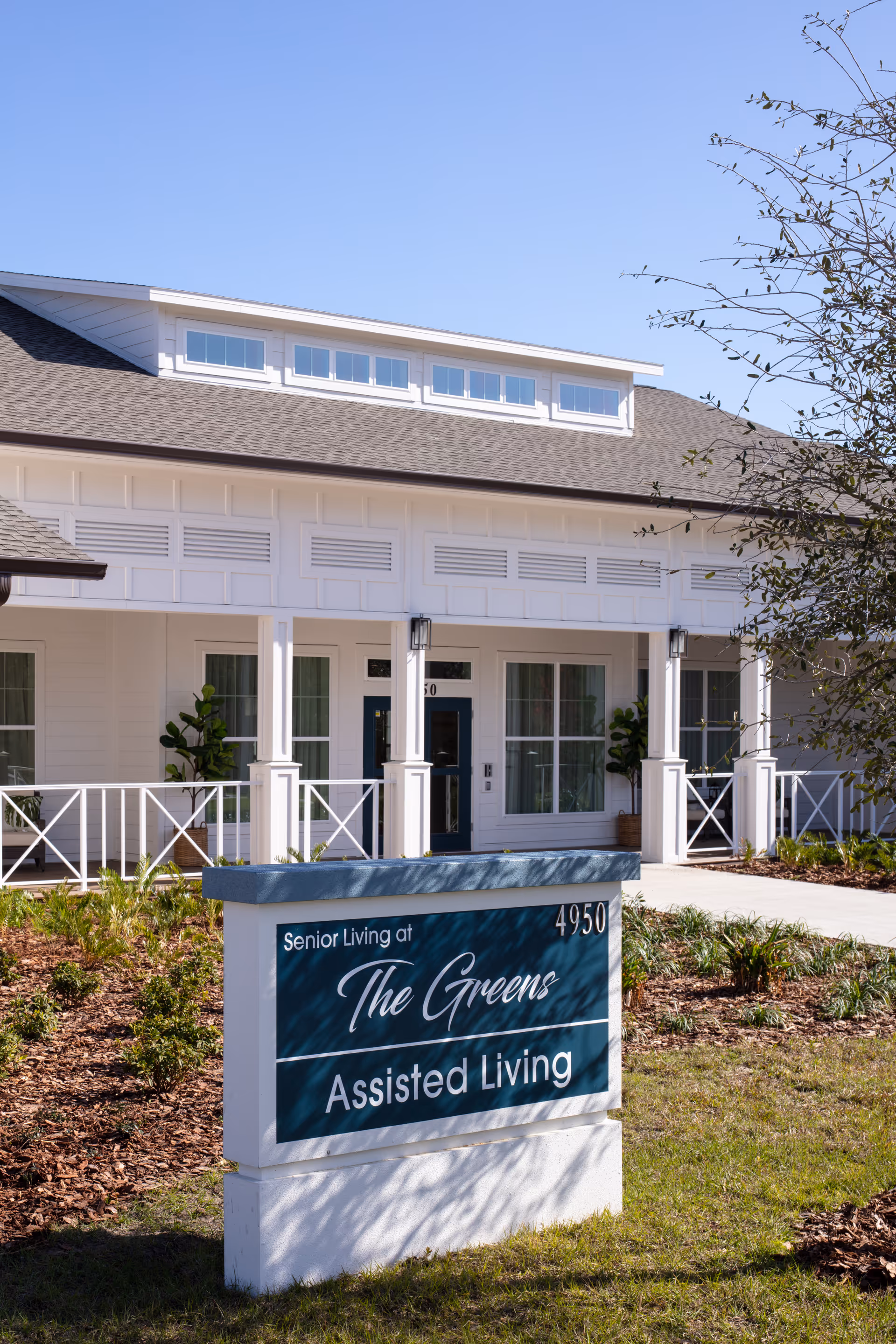 Exterior view of a senior living assisted living facility named Senior Living at The Greens, showing a white building with a porch, windows, and a sign in front with the facility name and address 4950.