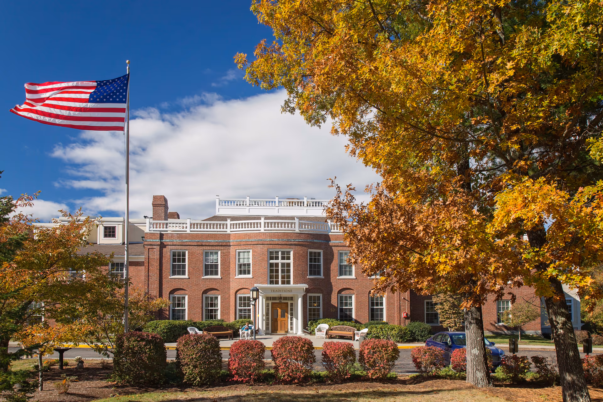Front exterior view of a large brick building with white trim, surrounded by trees with autumn foliage. An American flag is flying on a flagpole in front of the building. There are benches and shrubs near the entrance.
