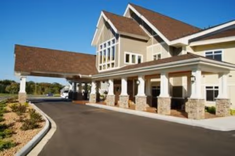 Exterior view of a senior living facility building with a covered entrance supported by stone pillars, a paved driveway, and landscaped areas with small plants under a clear blue sky.