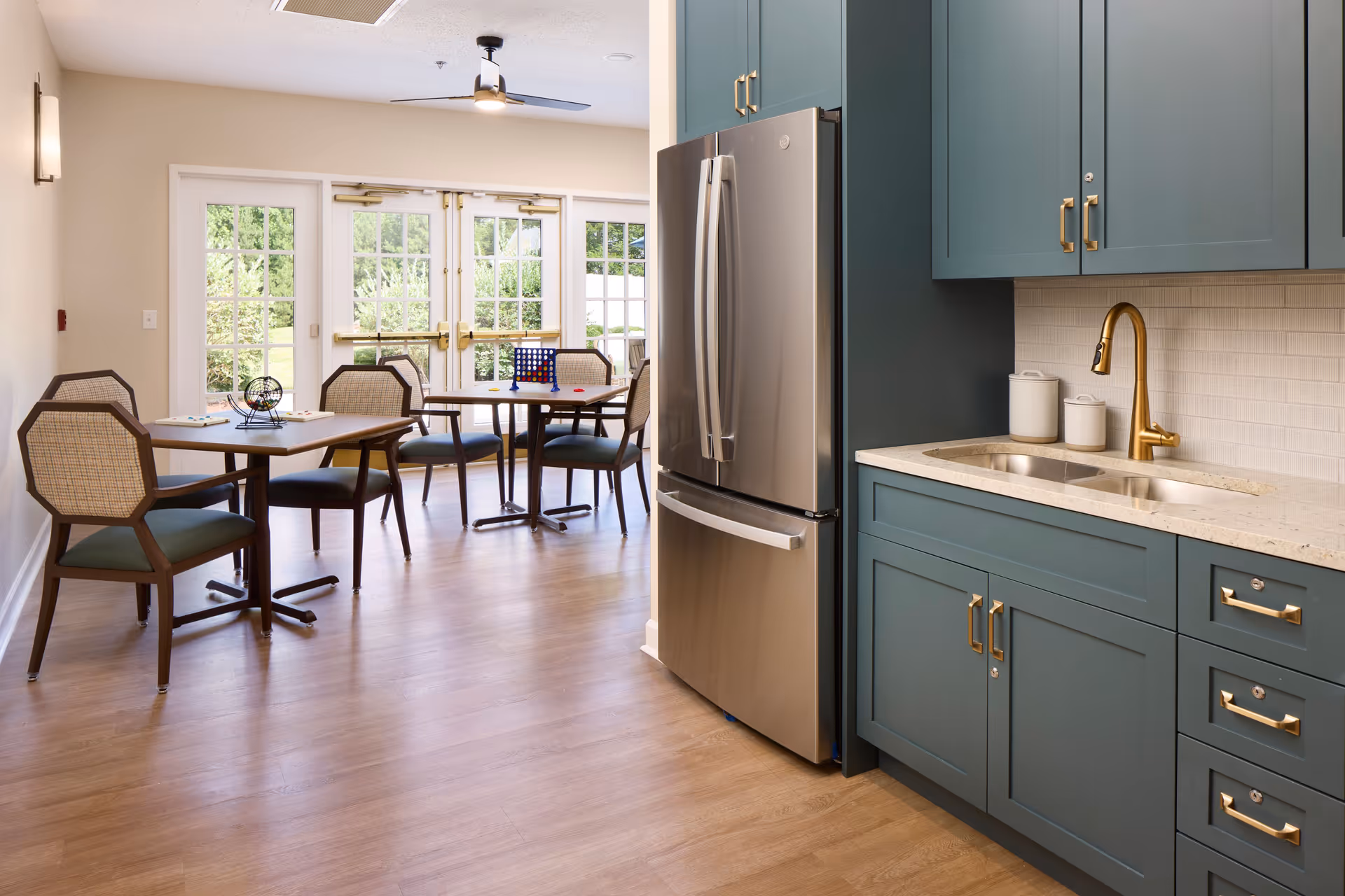 A bright room featuring a modern kitchen area with teal cabinets, a stainless steel refrigerator, and a gold faucet over a double sink. Adjacent to the kitchen are two tables with chairs, each set with a game, including bingo and Connect Four. Large glass doors and windows let in natural light and offer a view of greenery outside.