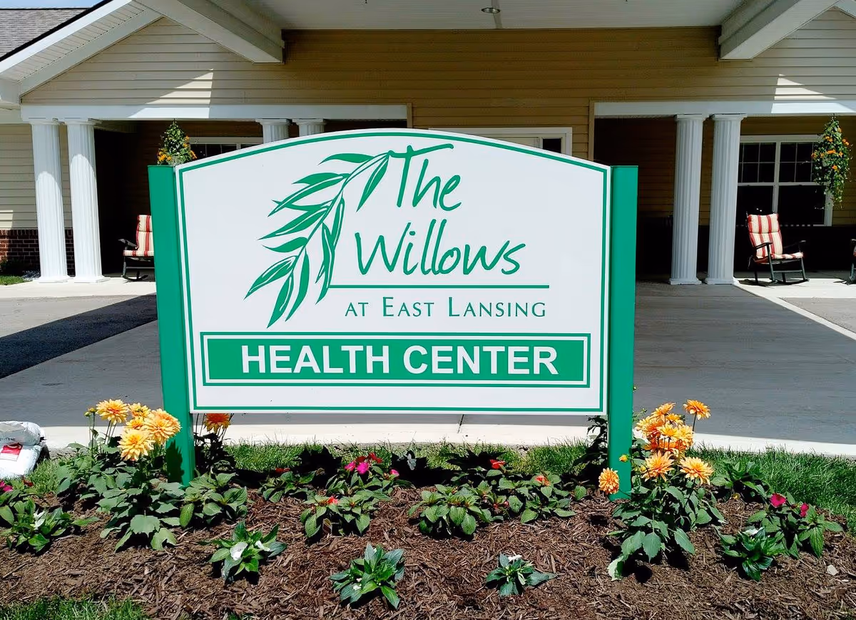 A green-and-white sign reading 'The Willows at East Lansing HEALTH CENTER' stands in a flowerbed in front of a building entrance with columns and chairs.