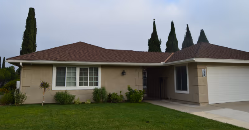 Single-story residential building with beige exterior walls, a brown shingle roof, and a well-maintained green lawn in front. There are several tall, narrow trees behind the building and a driveway leading to a white garage door on the right side.