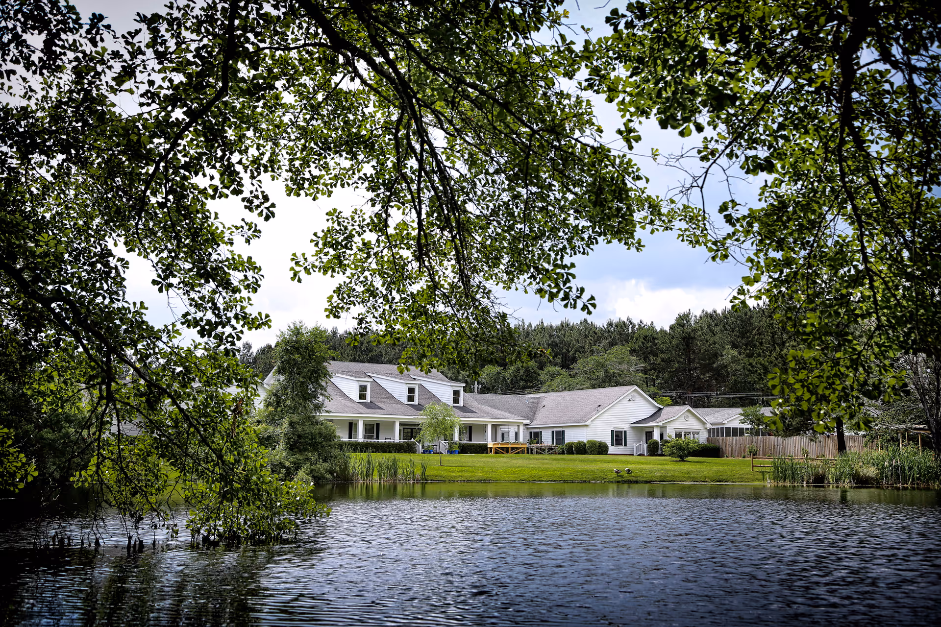 White senior care building with porches sits across a pond, framed by overhanging tree branches.