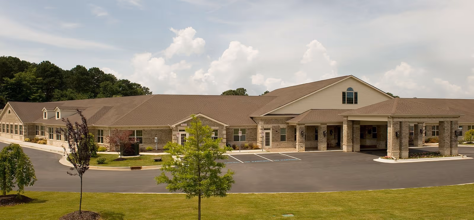 Exterior view of a single-story senior living facility building with a covered entrance, multiple windows, a paved driveway, and landscaped green lawn with small trees under a partly cloudy sky.