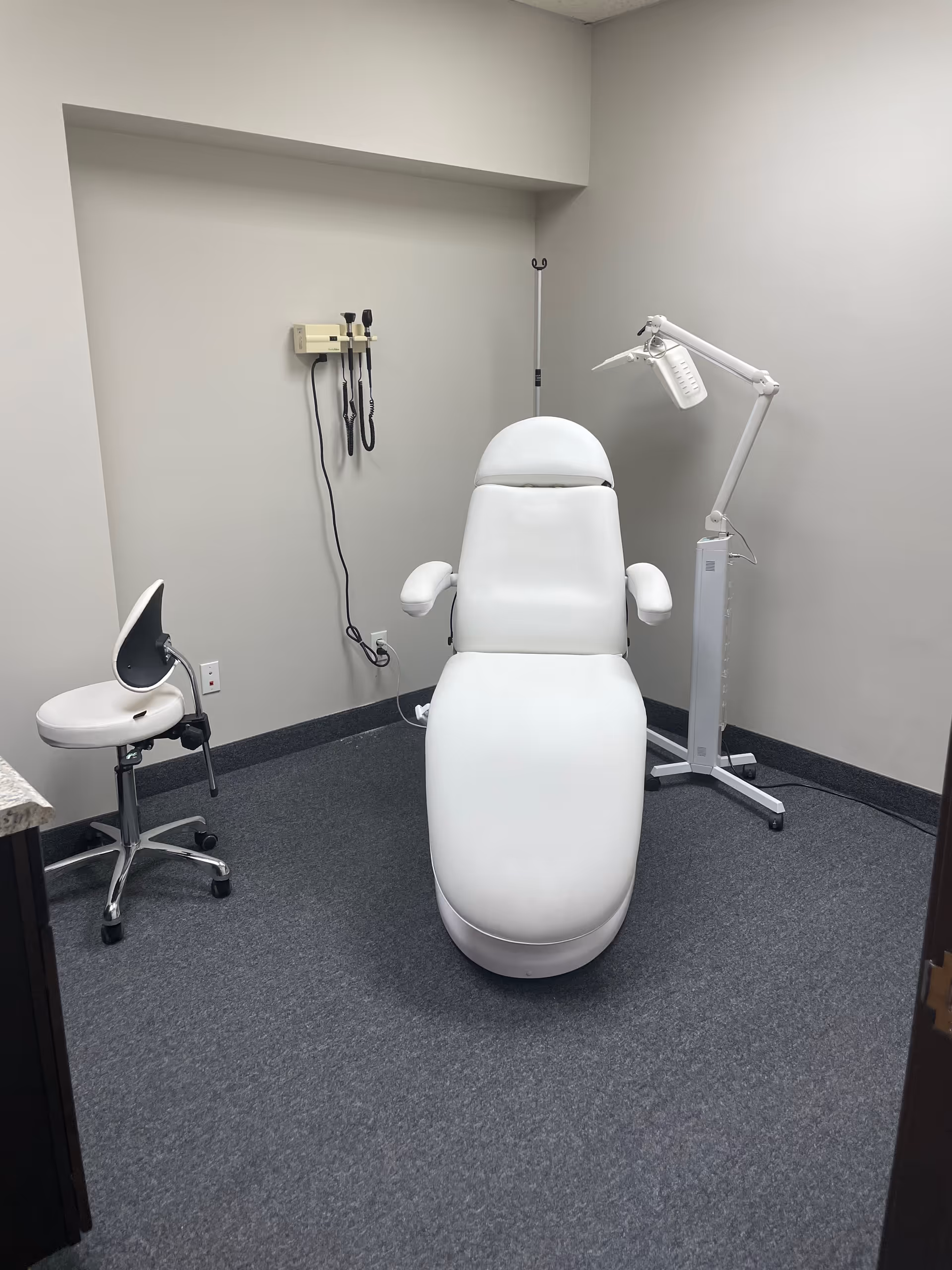 A white medical examination chair with a lamp and stool in a small clinical room.