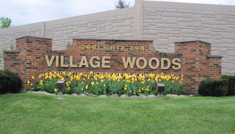 Exterior view of Village Woods sign with brick backdrop and yellow tulips in front.