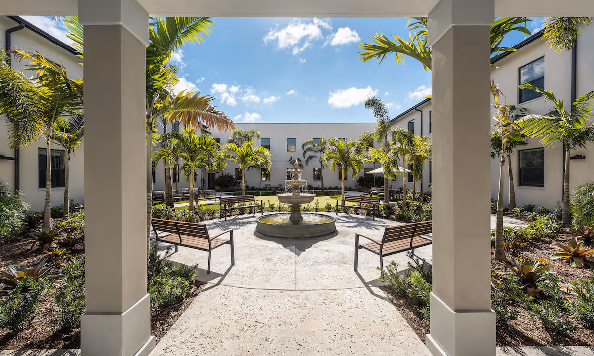 View of a sunny courtyard with a central water fountain surrounded by benches and palm trees, framed by two large pillars and a covered walkway. The courtyard is enclosed by a two-story building with multiple windows.