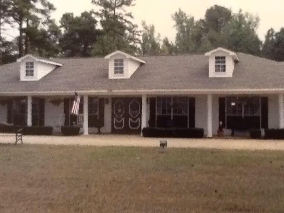 Front exterior view of a single-story building with a gray roof, three dormer windows, white columns supporting a covered porch, and a double front door. There is an American flag hanging on the porch and some bushes along the front of the building. The building is surrounded by trees and a grassy lawn.