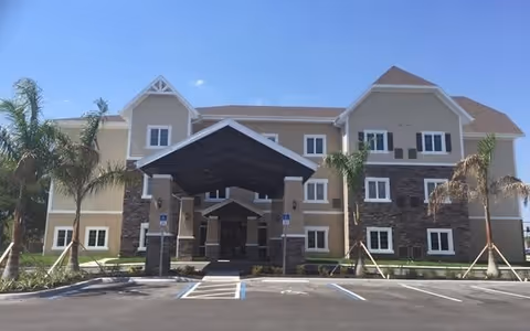 Front exterior view of a three-story assisted living facility building with beige and stone facade, a covered entrance, palm trees, and empty parking spaces in front under a clear blue sky.