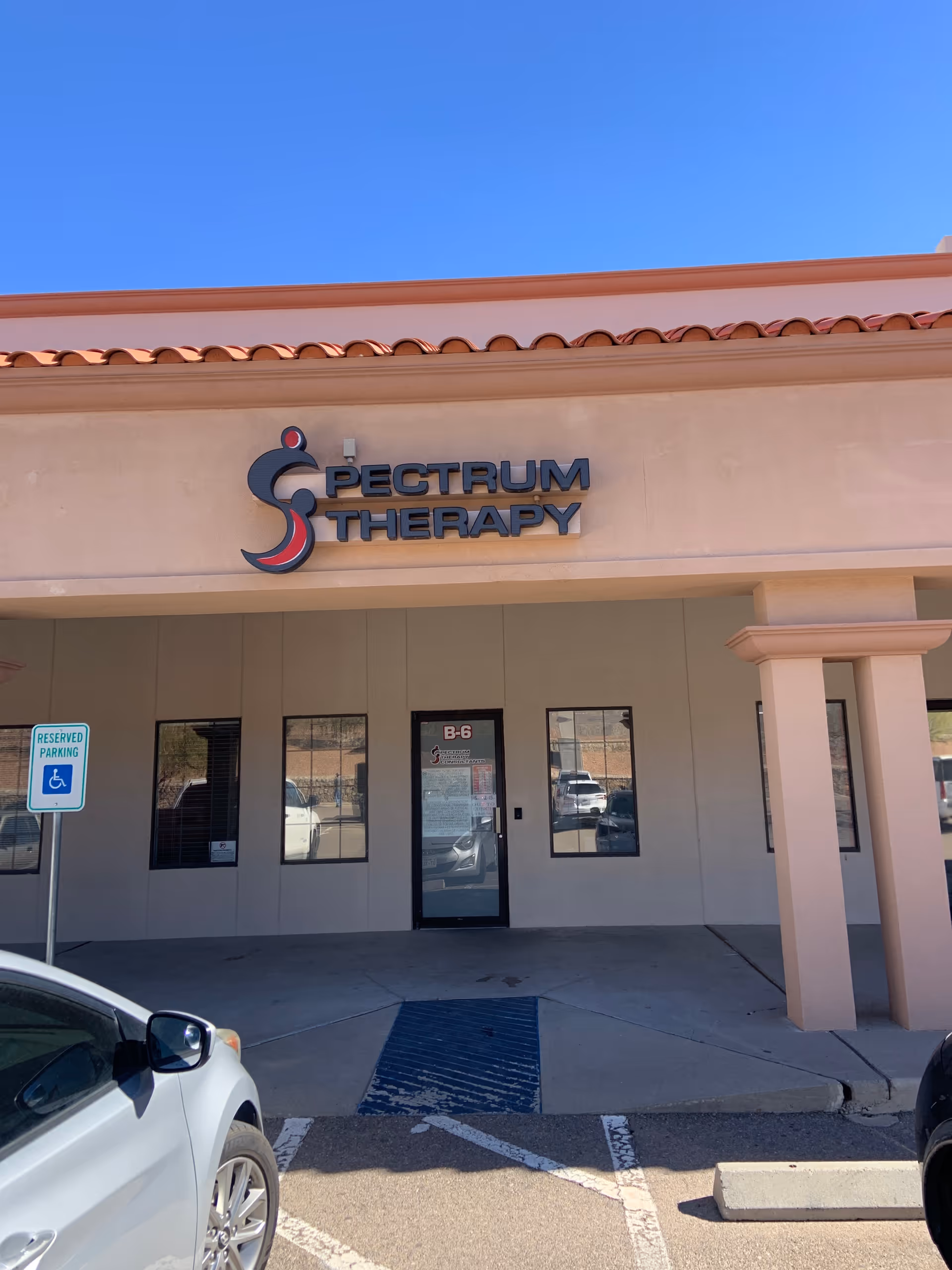 Front view of Spectrum Therapy facility with sign, windows, parked cars, and clear blue sky.
