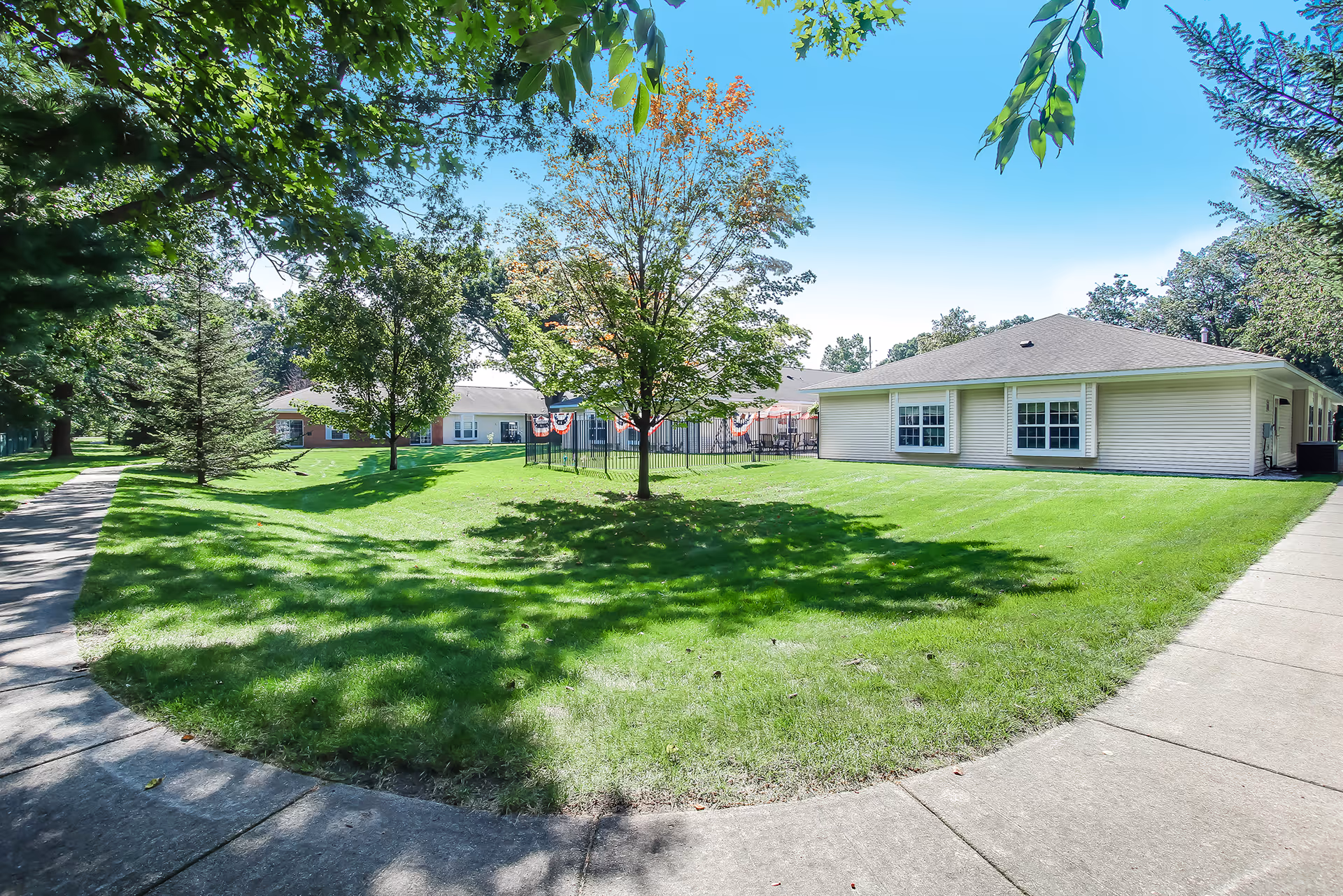 Outdoor view of Lakeview Terrace Assisted Living facility showing a well-maintained grassy area with trees and a curved concrete walkway. The single-story building with light-colored siding and multiple windows is visible on the right side. The sky is clear and blue, and there are some decorative banners on a fenced area in the background.