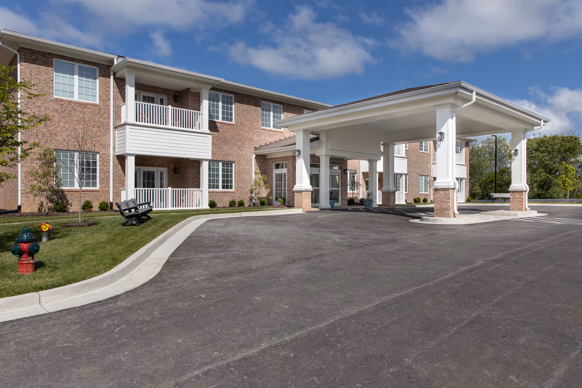Brick two-story senior living building with a covered porte-cochere entrance, balconies, landscaped lawn and paved driveway under a blue sky.
