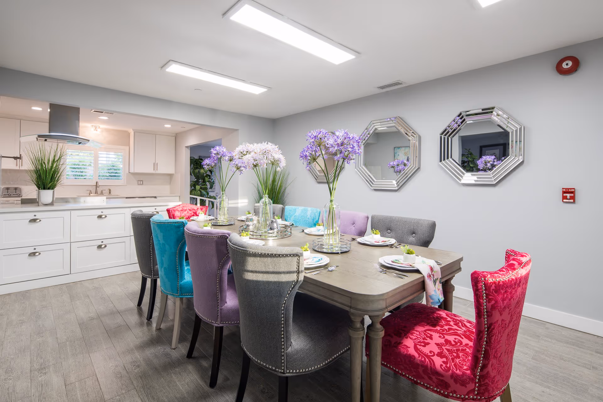 Dining area with a wooden table set with plates and vases of purple flowers surrounded by colorful upholstered chairs, with a white kitchen visible in the background.