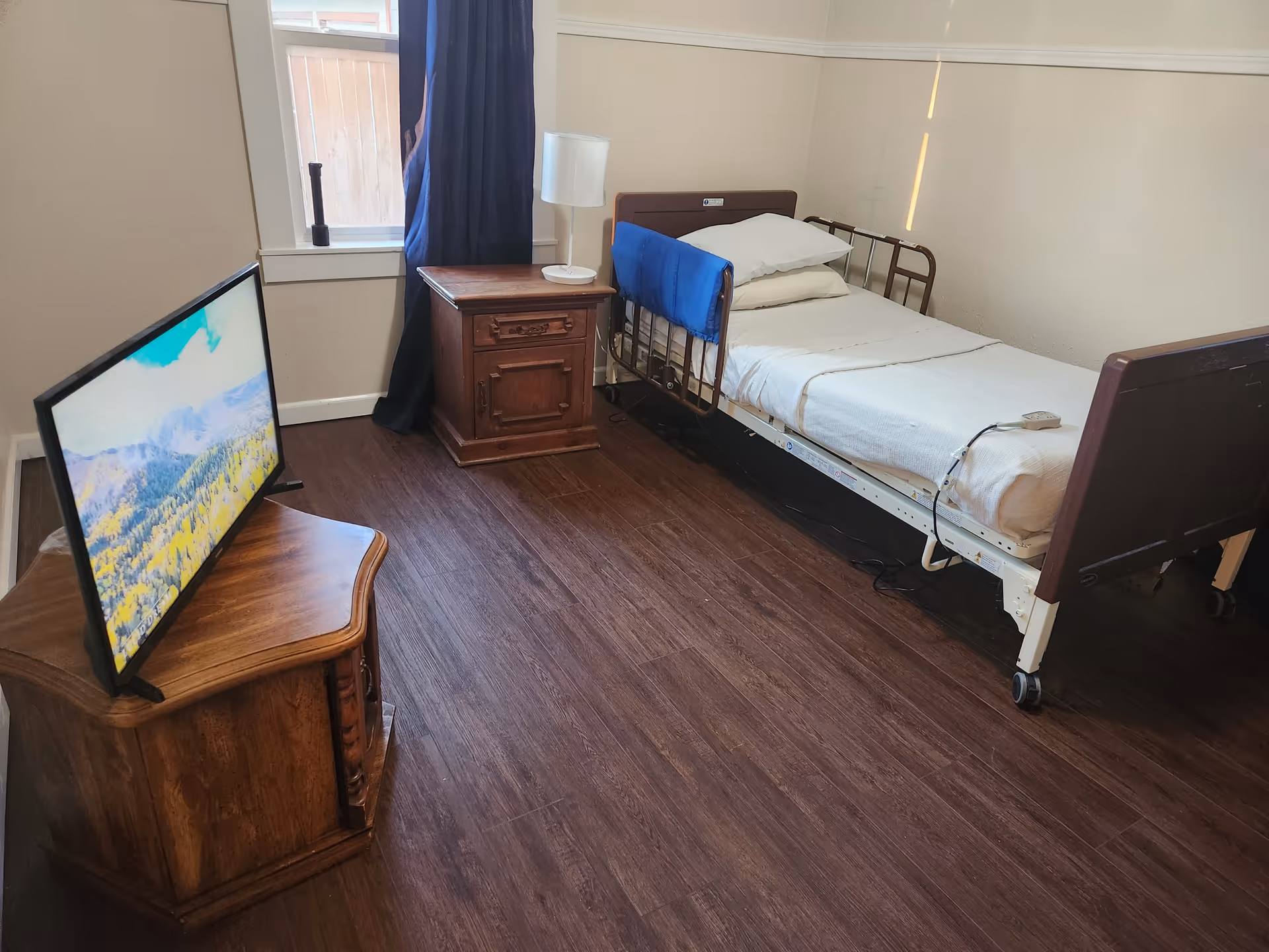 A hospital-style bedroom with a single bed, TV stand, nightstand, and window.