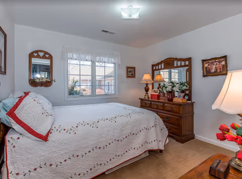 Cozy bedroom with a bed, dresser, mirrors, lamps, and a window at Elderberry Health Care.