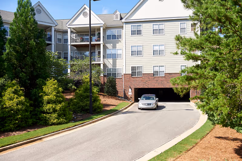 Front exterior of a multi-story senior living building with a driveway, parking garage entrance, and landscaped trees.