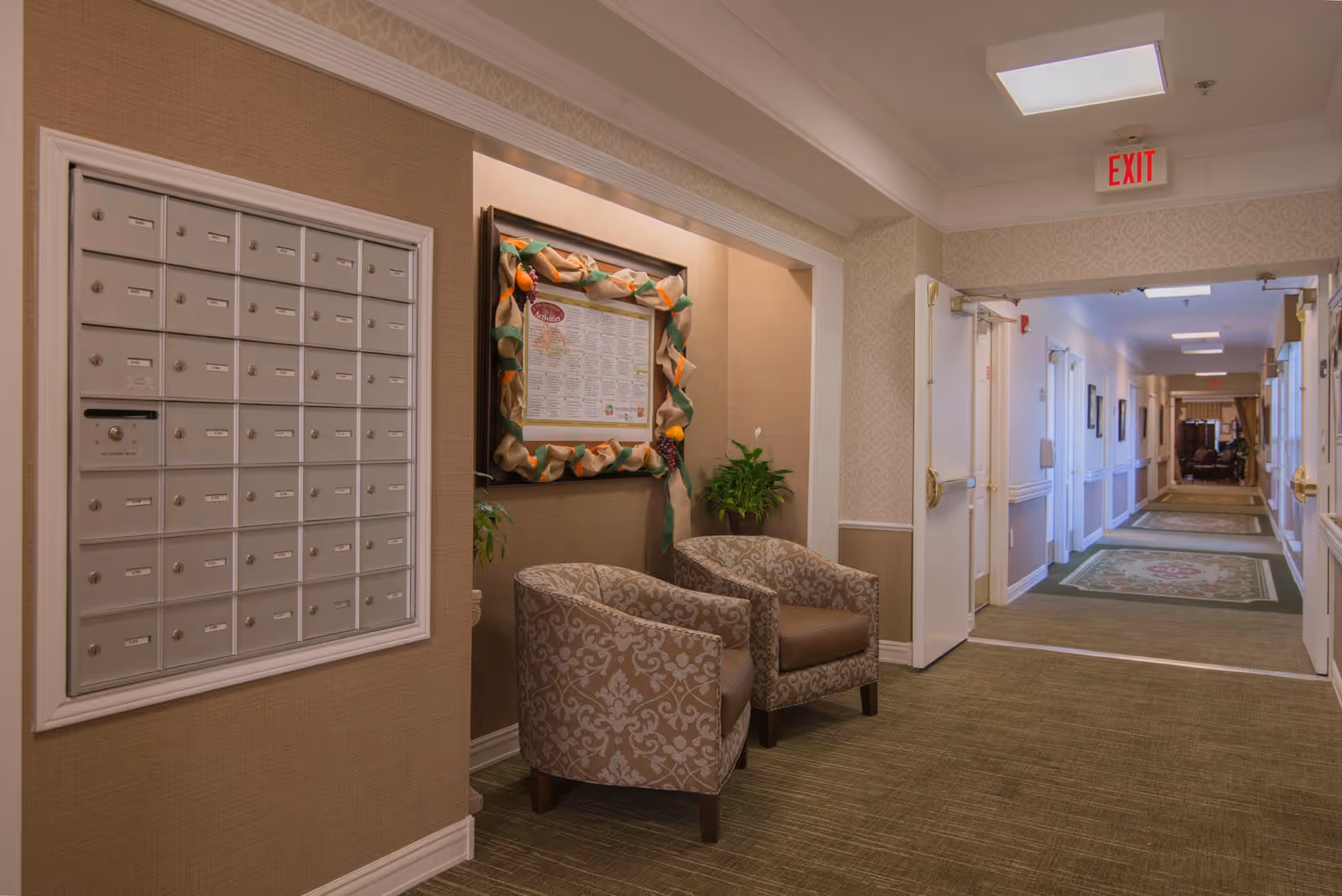 Carpeted interior hallway of a senior living facility with a bank of mailboxes, two upholstered chairs, a bulletin board, and a long corridor.