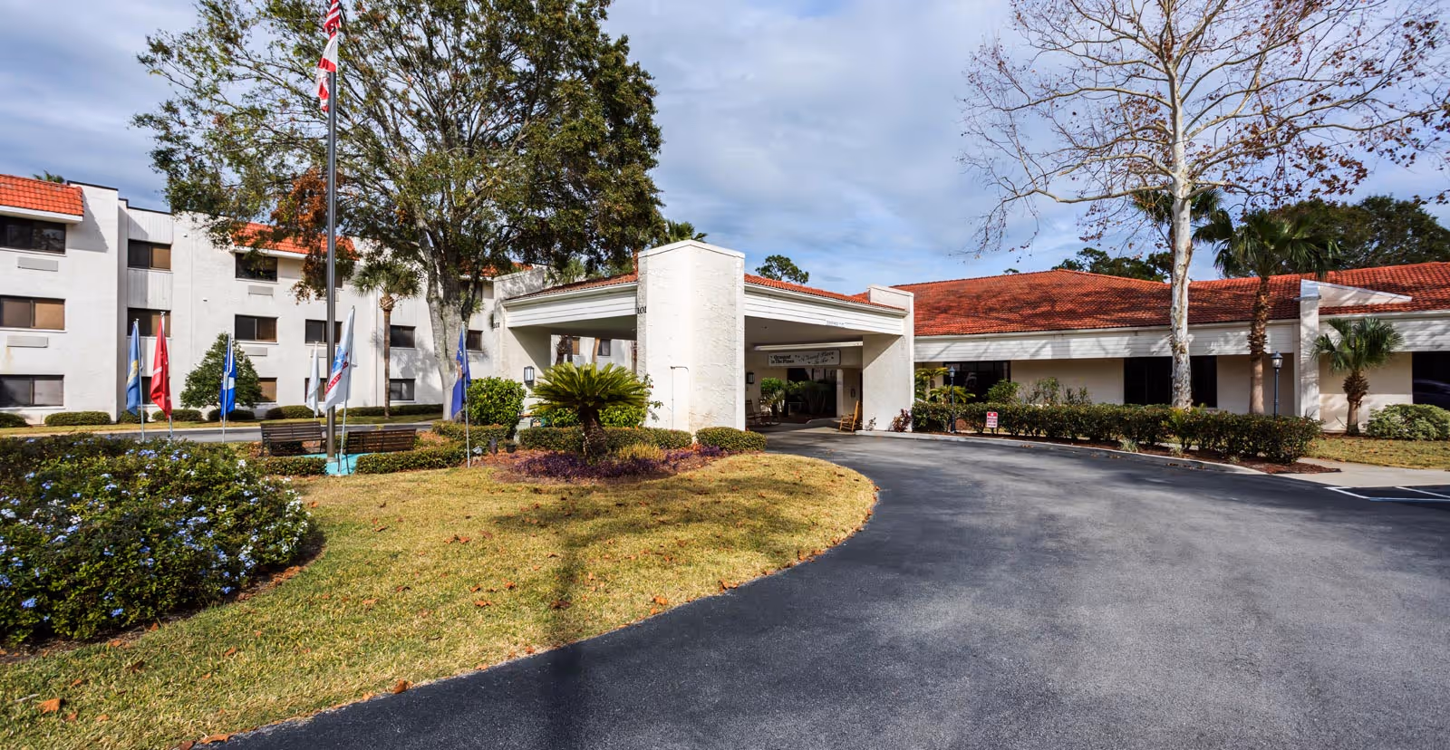 Front exterior of a senior living facility with a covered vehicle drop-off, flags, red-tiled roofs, and landscaped grounds.